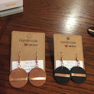 May include: Two pairs of handmade leather earrings with gold accents. The earrings are on brown card stock with the text "Handmade with love" printed on each card.