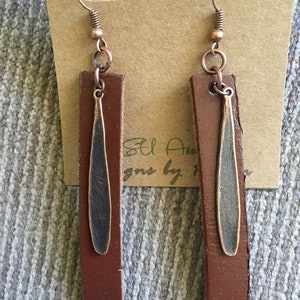 May include: A pair of brown leather earrings with a copper-toned metal pendant. The earrings are hanging from a brown card with the text "St. Anne Designs by" on it.