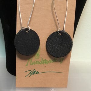 May include: A pair of black leather earrings with silver hooks. The earrings are round and have a textured surface. The earrings are handmade and are displayed on a brown card with the text "Handmade by Kas" written in green.