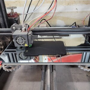 以下が含まれることがあります： ガラス製のベッドに黒いフィラメントが印刷されている3Dプリンターのクローズアップ。プリンターは黒と銀のフレームと赤と黒のノズルを持っています。