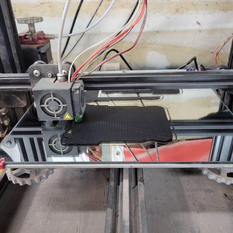 以下が含まれることがあります： ガラス製のベッドに黒いフィラメントが印刷されている3Dプリンターのクローズアップ。プリンターは黒と銀のフレームと赤と黒のノズルを持っています。