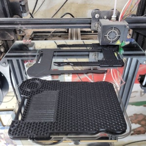 以下が含まれることがあります： 3Dプリンターが、ハニカム模様の黒いプラスチック製のオブジェクトを印刷しています。オブジェクトはガラス製のプラットフォームに印刷されています。