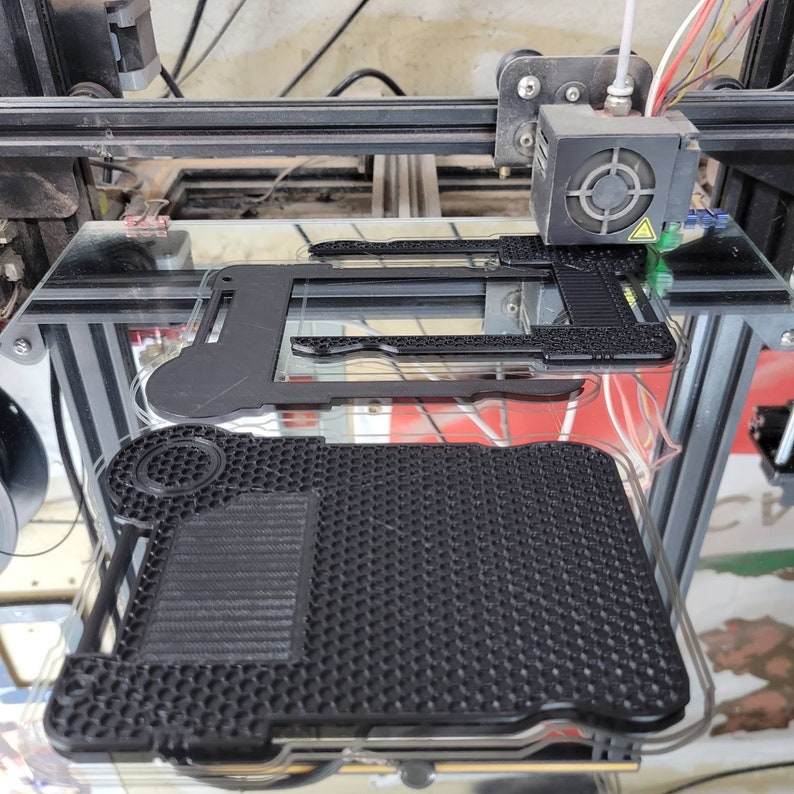 以下が含まれることがあります： 3Dプリンターが、ハニカム模様の黒いプラスチック製のオブジェクトを印刷しています。オブジェクトはガラス製のプラットフォームに印刷されています。