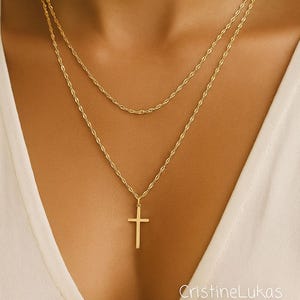 May include: A gold-colored, layered necklace with a cross pendant. The necklace features two delicate chains, one with a cross pendant and the other a simple chain. The jewelry is displayed against a white background.