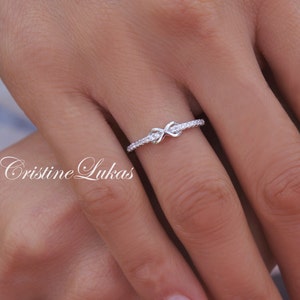 May include: A silver ring with an infinity knot design, encrusted with small, clear gemstones. The ring is on a finger, with the brand name "Cristine Lukas" in cursive script in the lower left corner. The ring is a delicate piece of jewelry.