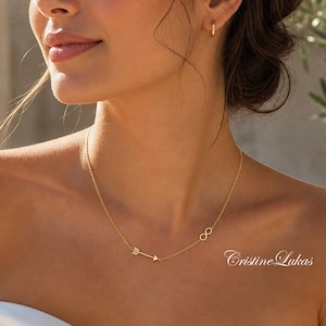 May include: A delicate gold necklace featuring an arrow and an infinity symbol. The necklace is worn on a person with a white strapless top. The brand name "Cristine Lukas" is visible in the image.