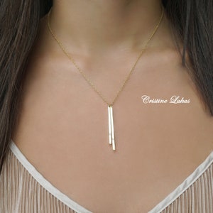 May include: A gold chain necklace with a pendant featuring two vertical bars with small diamonds. The necklace is worn by a person with long dark hair. The text "Cristine Lukas" is visible in the background.