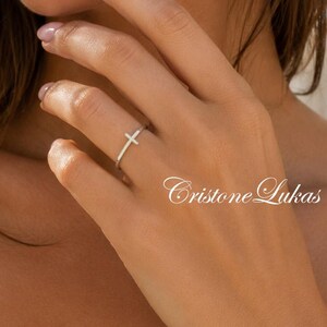 May include: A silver ring with a cross design. The ring is worn on a finger. The image also includes the text "Cristone Lukas".