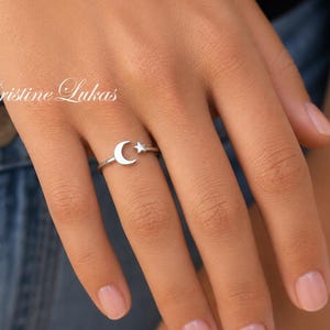 May include: A silver-toned ring featuring a small crescent moon and a tiny star motif on a thin band. The text Cristine Lukas is visible in white script lettering positioned above the ring. The ring is worn on a finger, showcasing the metal design.