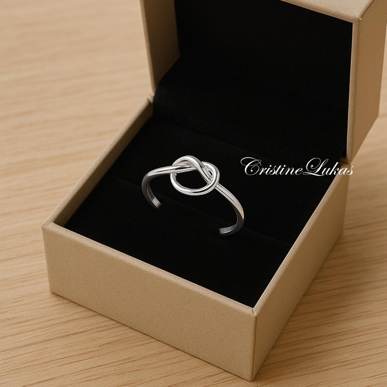 May include: A silver-colored ring with a knot design, presented in a beige jewelry box with a black interior. The ring is the focal point, showcasing a polished finish. The box is open, revealing the ring's intricate knot detail. The text "Cristine Lukas" is visible.