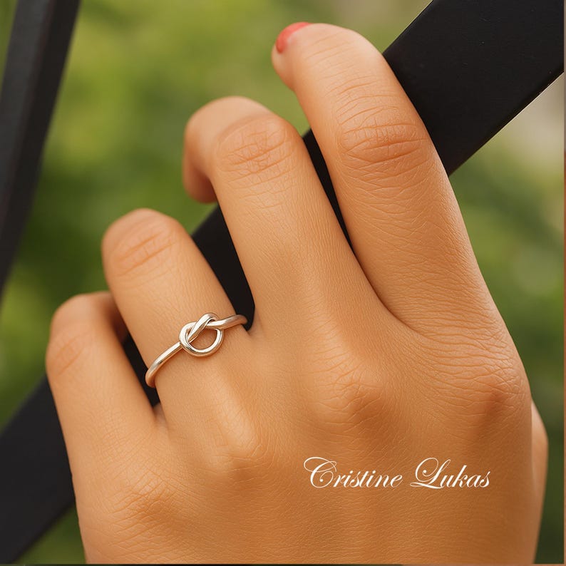 May include: A silver-colored knot ring is worn on a finger. The ring is the focal point, with a simple knot design. The background is blurred, with a green and brown color scheme. The text "Cristine Lukas" is in the bottom right corner.