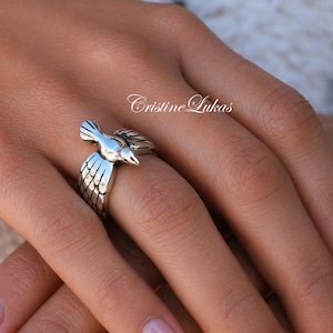 May include: A silver-toned ring featuring a detailed bird design. The bird is depicted in flight, with outstretched wings and a detailed feather pattern. The ring is a unique piece of jewelry with the text "Cristine Lukas".