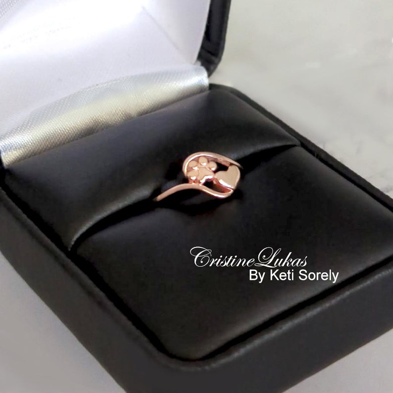 May include: Rose gold ring with a paw print and heart design. The ring is in a black ring box with the text "Cristine Lukas By Keti Sorely".