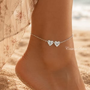 May include: A silver anklet featuring a delicate chain and two small heart-shaped charms. Each heart charm is engraved with a single letter, specifically the letters S and W. The jewelry piece is displayed on an ankle against a beach background.