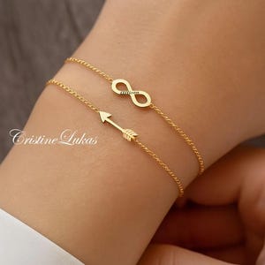 Puede incluir: Se muestran dos delicadas pulseras doradas en una muñeca. La pulsera superior presenta un símbolo de infinito con el nombre "allison" inscrito. La pulsera inferior tiene un diseño de flecha. Las pulseras están conectadas por una fina cadena.