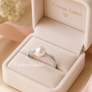 May include: A pearl ring in a velvet jewelry box. The ring has a large, round pearl and a silver band with small, sparkling stones. The box is beige and has the name "Cristine Lukas" printed on the inside.