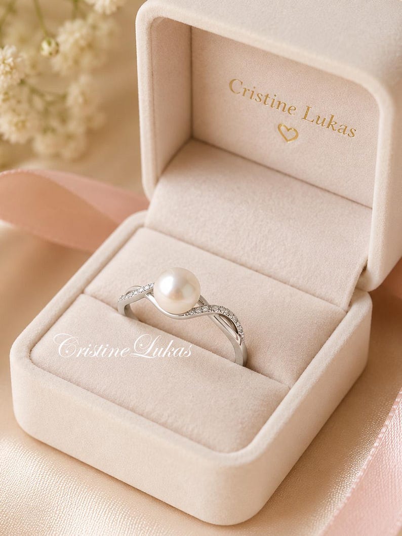 May include: A pearl ring in a velvet jewelry box. The ring has a large, round pearl and a silver band with small, sparkling stones. The box is beige and has the name "Cristine Lukas" printed on the inside.
