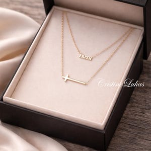 Peut inclure: Un ensemble de colliers dorés dans un coffret noir. L'ensemble comprend deux colliers, l'un avec le mot "FAITH" et l'autre avec un pendentif en forme de croix latérale. La boîte est doublée d'un tissu beige clair.