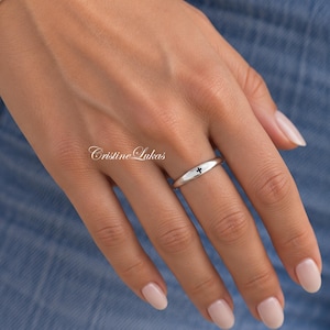 May include: A polished silver band ring featuring a small, centered black cross symbol. The ring has a smooth, rounded profile and is worn on the ring finger. The text Cristine Lukas is visible in a cursive script font near the ring.
