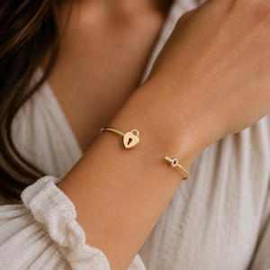 May include: A gold-coloured open cuff bracelet. One end features a heart-shaped padlock design, while the other end has a knot embellished with small, clear gemstones. The bracelet is worn on the wrist.