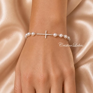 May include: A silver bracelet featuring a cross pendant and a string of white pearls. The bracelet is displayed on a hand against a soft, neutral-toned background. The text "Cristine Lukas" is visible.