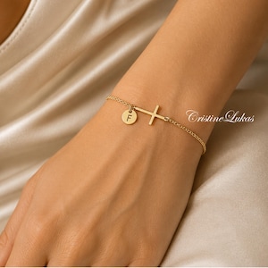 Op de afbeelding: Een goudkleurige kettingarmband met een kleine ronde bedel gegraveerd met de letter F en een zijwaartse kruisbedel. De armband heeft een delicaat en minimalistisch ontwerp, met de merknaam Cristine Lukas zichtbaar in de afbeelding.