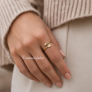 May include: A gold-colored arrow ring with two arrowheads, one pointing up and the other down. The ring wraps around the finger. The ring is on a hand, with the text "Cristine Lukas" in cursive at the bottom.