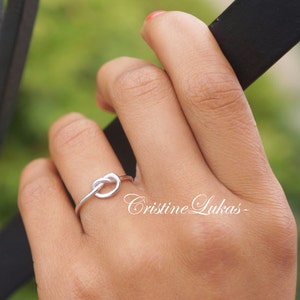 May include: A silver-colored knot ring. The ring is a simple design, with the knot as the focal point. The ring is on a finger, with a blurred green background. The text "Cristine Lukas" is in the image.