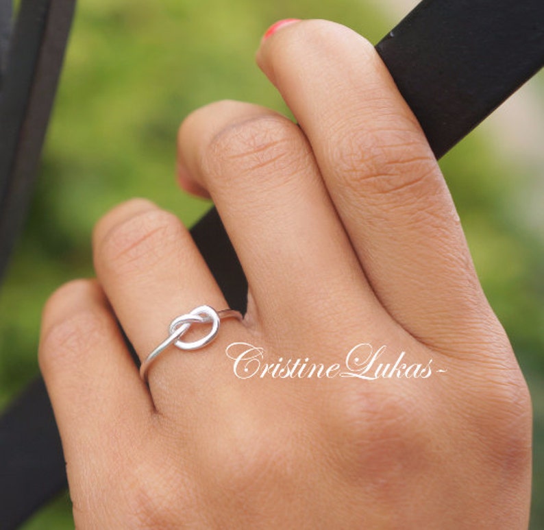May include: A silver-colored knot ring. The ring is a simple design, with the knot as the focal point. The ring is on a finger, with a blurred green background. The text "Cristine Lukas" is in the image.