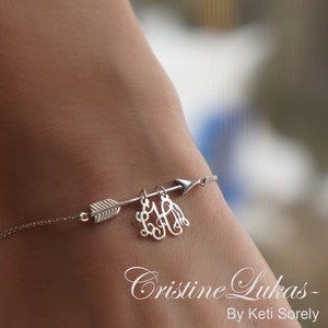 May include: A silver chain bracelet with a silver arrow charm and a silver monogram charm. The monogram charm is a cursive "LHA".