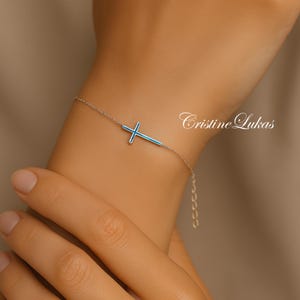 May include: A silver bracelet with a cross pendant, filled with blue enamel. The bracelet is displayed on a wrist. The brand name "Cristine Lukas" is visible in the background.