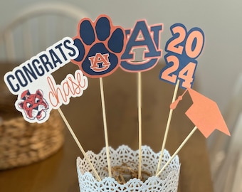 May include: A white metal vase with a lace design, holding a graduation centrepiece with orange and blue decorations. The decorations include a paw print with the letters "AU", a graduation cap, the letters "A" and "2024", and a tiger mascot with the words "Congrats chase".