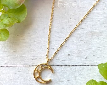 Könnte beinhalten: Eine goldene Kettenkette mit einem Halbmond-Anhänger, der kleine Sterne im Inneren des Mondes trägt. Die Kette liegt auf einer weißen Holzoberfläche.
