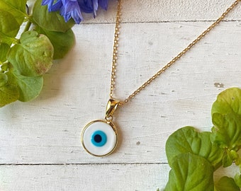 Puede incluir: Un collar de cadena dorada con un colgante de ojo turco blanco y azul. El colgante es redondo y tiene un ojo azul con una pupilla blanca y un iris negro. El collar está sobre una superficie de madera blanca con hojas verdes y una flor azul.