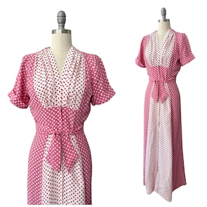 May include: A vintage pink and white polka dot dress with short sleeves and a matching belt. The dress is displayed on a mannequin and features a V-neckline and a button-down front. The fabric has a repeating pattern of small white dots.