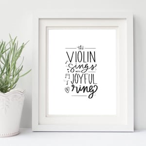 May include: A framed print with the words "the VIOLIN sings with JOYFUL ring" in black script. The print is in a white frame, and a small potted plant sits to the left. The image has a clean, minimalist aesthetic.