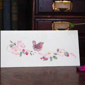 May include: Watercolor illustration of a small brown bird perched on a branch with pink flowers and berries. The artwork is on a rectangular, off-white card, displayed on a wooden surface.