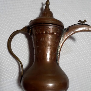 May include: A copper-colored, antique-style coffee pot with a curved handle and a spout. The pot features intricate etched designs and a decorative bird-shaped finial on the lid. The pot is likely made of copper or a copper alloy.