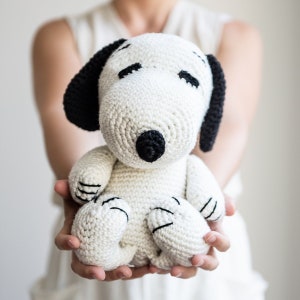 May include: A white crocheted stuffed dog with black ears and a black nose. The dog is sitting in the hands of a person.