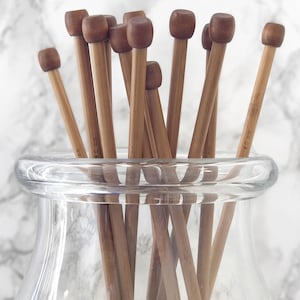 May include: A glass jar filled with a bundle of natural wooden knitting needles. The needles are various sizes and are labeled with their size in millimeters.
