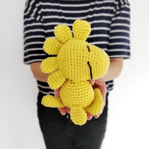 May include: A yellow crocheted stuffed animal shaped like a dinosaur with black eyes. The dinosaur is being held by a person wearing a striped shirt.