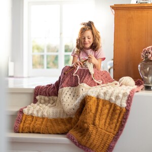 May include: A young person sits on a white step, crocheting a multi-colored blanket with a chunky yarn. The blanket is made of different colored stripes, including cream, mustard yellow, and pink.