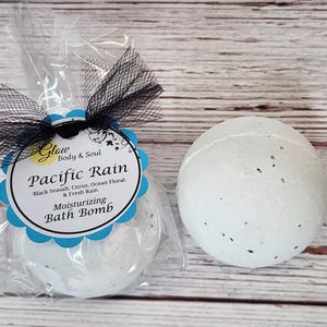 May include: A white bath bomb with black specks, packaged in a clear plastic bag with a black and white tulle bow. The bath bomb has a blue and white label that reads "Glow Body & Soul Pacific Rain Black Seasalt, Citrus, Ocean Floral & Fresh Rain Moisturizing Bath Bomb".