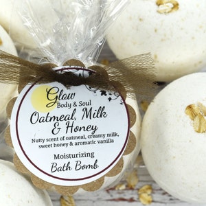 May include: Four white bath bombs with oatmeal on top, wrapped in clear plastic with a brown ribbon and a label that reads "Glow Body & Soul Oatmeal, Milk & Honey Nutty scent of oatmeal, creamy milk, sweet honey & aromatic vanilla Moisturizing Bath Bomb"