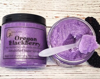 Op de afbeelding: Een glazen pot met paarse suiker scrub met een zwarte deksel. Het etiket vermeldt "Oregon Blackberry" en "Emulsified Sugar Scrub". De scrub is gemaakt met rijpe bramen, vanille en groen. Gemaakt met biologische en pure rietsuiker.