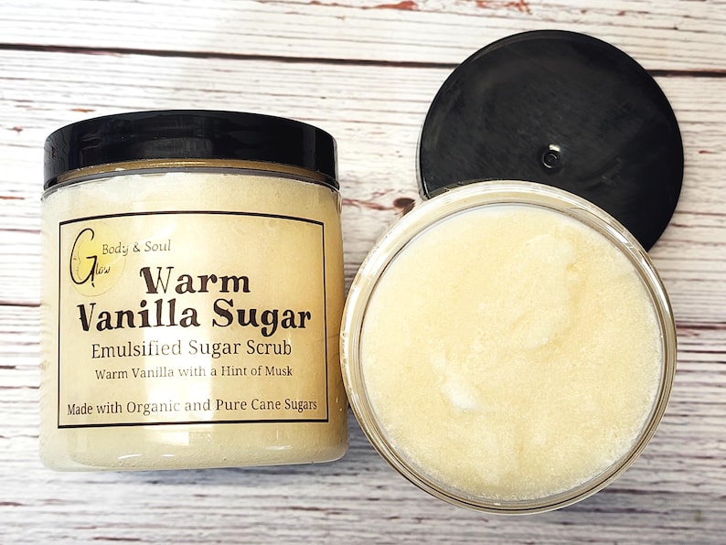 Op de afbeelding: Een glazen pot met warme vanille-suikerscrub. De pot heeft een zwarte deksel en een wit etiket met de tekst "G Body & Soul Glow Warm Vanilla Sugar Emulsified Sugar Scrub Warm Vanilla with a Hint of Musk Made with Organic and Pure Cane Sugars".