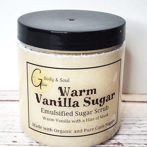 Op de afbeelding: Een glazen pot met een zwarte deksel gevuld met een lichtbruine suiker scrub. Het etiket luidt "Body & Soul Glow Warm Vanilla Sugar Emulsified Sugar Scrub Warm Vanilla with a Hint of Musk Made with Organic and Pure Cane Sugars".