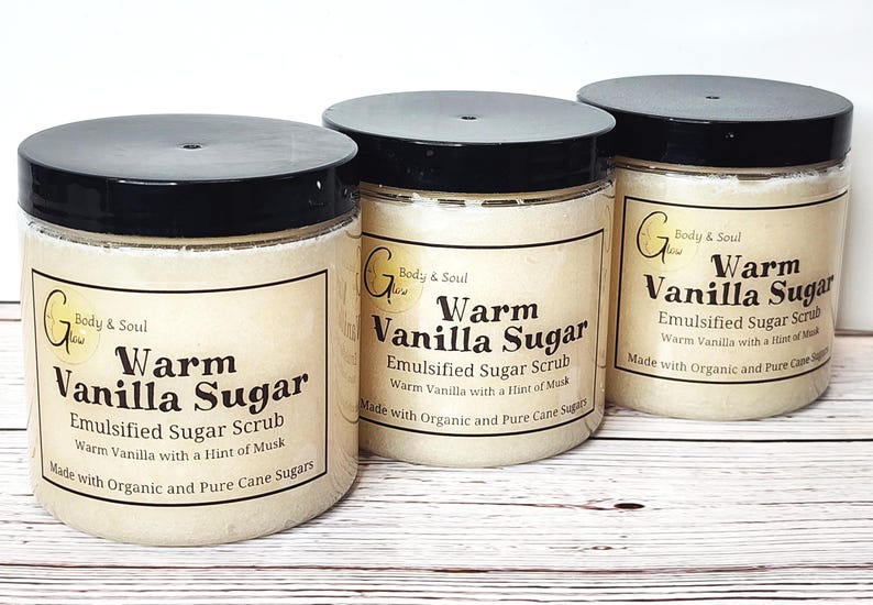 Op de afbeelding: Drie glazen potten gevuld met een lichtbruine suiker scrub. Elke pot heeft een zwarte deksel en een wit etiket met de tekst "Warm Vanilla Sugar" en "Emulsified Sugar Scrub". Het etiket bevat ook de tekst "Warm Vanilla with a Hint of Musk" en "Made with Organic and Pure Cane Sugars".