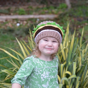 May include: A young child wearing a green and white patterned dress and a brown, white, and green crocheted hat shaped like a hamburger.