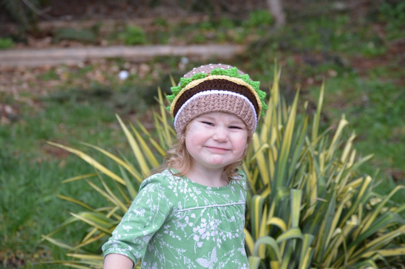 May include: A young child wearing a green and white patterned dress and a brown, white, and green crocheted hat shaped like a hamburger.
